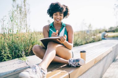 Gülümseyen, siyah saçlı, kıvırcık saçlı, planlamacıyla oturan, açık havada fikirlere dikkat çeken, mutlu, Afro-Amerikan hipster kız blog için yayın metni yazan, yedek kulübesinde eğlenen.