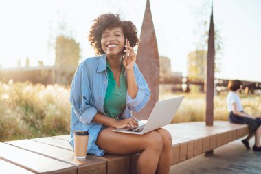 Kıvırcık saçlı, dizüstü bilgisayarda yazan ve cep telefonuyla konuşan neşeli Afro-Amerikan hippi kız portresi. Parkta uzaktan kumandayla eğlenen mutlu, esmer tenli bir kadın öğrenci sokakta cihaz kullanıyor. 