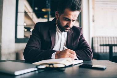 Düşünceli, beyaz sakallı bir adam not defteri planlama programına yazıyor. Ofisinde oturuyor, resmi giyinmiş düşünceli bir erkek girişimci iş süreci finans stratejilerini düzenleyen notlar yazıyor.