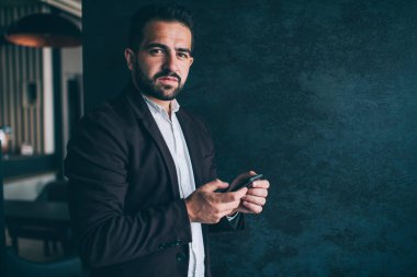 Başarılı, kendine güvenen iş adamının yarım boy portresi. Resmi moda kıyafetleriyle duvarın yanında duruyor. Akıllı telefonlu yakışıklı erkek CEO kameraya bakarak akıllı telefonuyla poz veriyor.