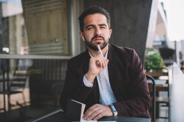 Resmi olarak giyinmiş ciddi bir hipster 'ın portresi kameraya bakıp kafeterya terasında oturup hayal ürünü beyaz iş adamının çalışma saatlerini not alarak yayınlama fikri üzerine kafa yoruyor.