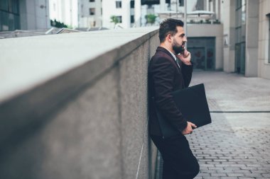 Resmi giysiler içinde kendine güvenen beyaz erkek girişimci ve kağıt klasörlü cep telefonu konuşmaları şehir ortamlarında duruyor, genç iş adamı akıllı telefonlardan dışarıda vakit geçiriyor.