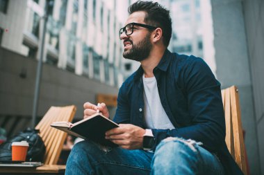 Görme düzeltmesi için göz giysisi giyen düşünceli beyaz erkek yazar açık havada defter yazma üzerine kafa yoruyor, düşünceli erkek öğrenci planlamacıyla birlikte oturup ev ödevlerini başka yöne çeviriyor.