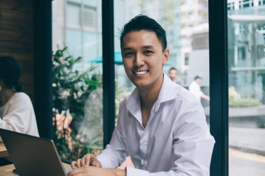 Beyaz gömlekli genç Asyalı adam gülümsüyor ve kafede dizüstü bilgisayarla beyaz masada oturmuş pencerelere bakan kameraya bakıyor.