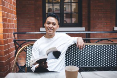 Neşeli modern Asyalı erkek okuyucu, kırmızı tuğla duvarlı kafede otururken komik bir kitap ve kahveyle vakit geçiriyor.