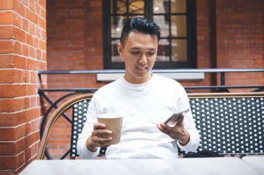 Neşeli genç Asyalı erkek öğrenci elinde akıllı telefon ve içecek tutarken dinleniyor ve sokak kafesinde masada oturuyor.