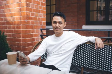 Tatmin olmuş Asyalı beyaz ceketli bir erkek öğrenci rahat açık hava kafeteryasında içki içerken cep telefonu kullanıyor ve kameraya bakıyor. 