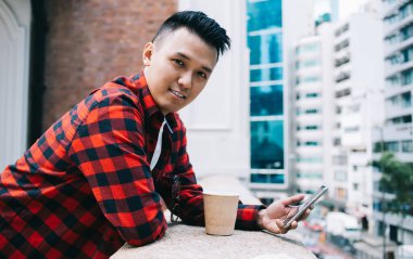 Kırmızı tişörtlü neşeli Asyalı bir erkek kameraya bakıyor, cep telefonunu tutuyor ve balkona yaslanıyor.