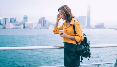 Düşünceli kadın turist, elinde kahveyle Hong Kong 'da nehir kenarında tatillerde kafa yoruyor. Çekici hippi kadın, Çin tatilleri sırasında metropolü keşfetmeyi düşünüyor.