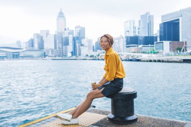 Moda kıyafetleri içinde mutlu hippi kız yaz günü şehir nehrinin kenarında dinleniyor günlük giysiler giymiş neşeli genç bayan turist arka planda Hong Kong 'la şehir ortamında gülüyor.