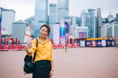 Sırt çantalı güzel Asyalı mutlu kadın fotoğraf çekiyor modern gökdelenli dijital akıllı telefon ve arka planda eğlence parkı kullanıyor.