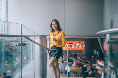 Neşeli, genç ve güzel Asyalı kadın, gündelik giysiler içinde kameraya bakarken ışıl ışıl gülümsüyor ve merdivenlerden akıllı telefon ve modern tasarım ile birlikte bir bardak içecekle çıkıyor.