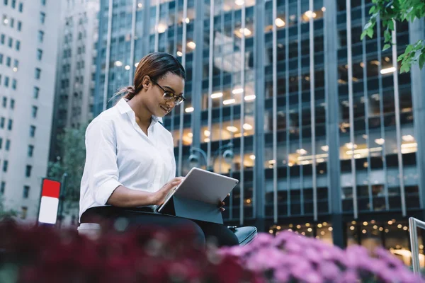 Young content African American spectacled female entrepreneur in corporate clothes using digital tablet while sitting in street during coffee break