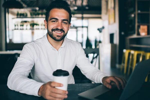 Portrait of cheerful caucasian man freelancer sitting in coffee shop with laptop computer working remotely satisfied with income, happy bearded male millennial have successful online business