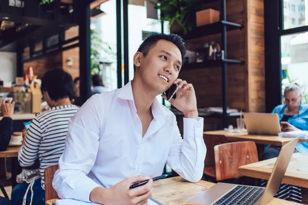 Gleeful and appealing Asian freelancer in neat shirt talking on phone and sitting in front of laptop in cozy cafe