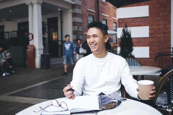 白いセーターを着た幸せな大人のアジアの男性は ノートパッドでテーブルに座ってカフェテラスでコーヒーを飲みに行くために保持しながら離れて見て笑っています — ストック写真