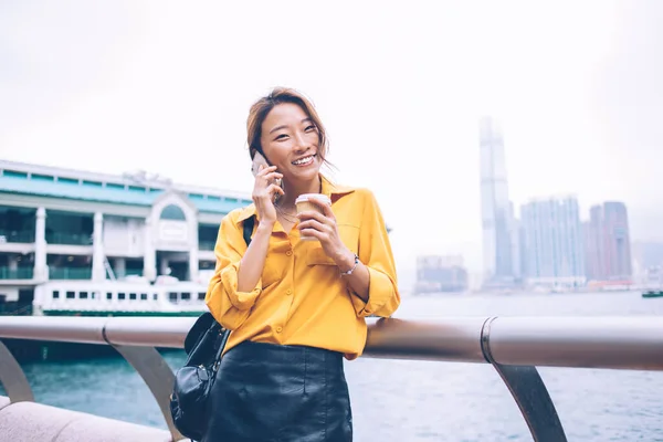 美しいアジアの女性は黄色のブラウスと黒の革のスカートを着て笑顔とフェリー乗り場の背景に香港市内でテイクアウトコーヒーを保持携帯電話を呼び出します — ストック写真
