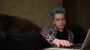 Genç adam evdeki kanepede dizüstü bilgisayar kullanıyor. Yazıyor, araklıyor. Adam evde dinleniyor.