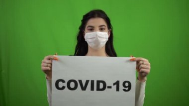 Tıbbi maskeli bir kız yeşil ekranda COVID-19 işareti tutuyor. Coronavirus şarkısı.