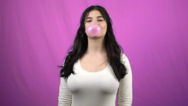 Morena chica soplando gran goma de mascar rosa sobre fondo púrpura en cámara lenta — Vídeo de stock