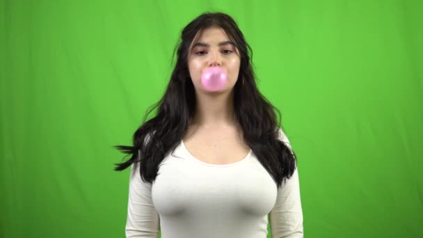 Jong brunette meisje op groen scherm blazen bubble gum en kijken naar camera — Stockvideo