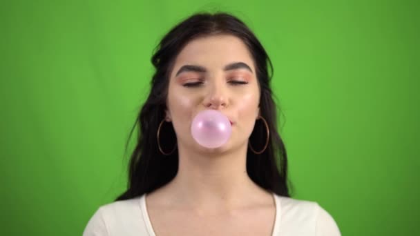 Funny smiling girl chewing bubble gum, blowing pink bubble gum on green screen — Stock Video