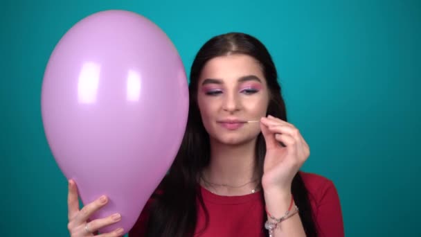 Jeune femme brune heureuse popping un ballon avec une aiguille sur fond bleu — Video