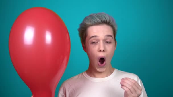 Joven feliz hombre con el pelo azul haciendo estallar un globo con aguja sobre fondo azul — Vídeos de Stock