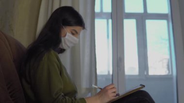Tıbbi maskeli kadın evde kalıp eskiz defterine resim çiziyor, karantina altında.