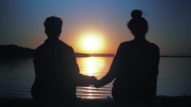 Para zakochana siedzi na plaży, patrzy na zachód słońca, relaksuje się, trzyma się za ręce — Wideo stockowe