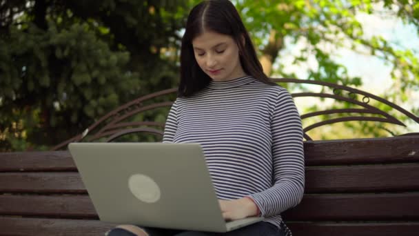 Junge Freiberuflerin arbeitet im Park, benutzt Laptop, sitzt auf Bank — Stockvideo