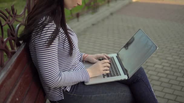 Νεαρή γυναίκα ελεύθερη επαγγελματίας που εργάζεται με φορητό υπολογιστή κάθεται στον πάγκο στο πάρκο της πόλης — Αρχείο Βίντεο