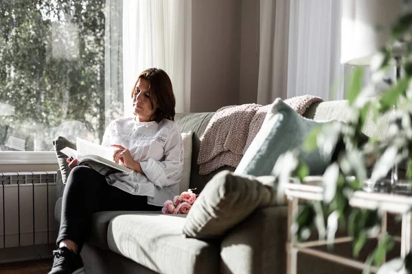 白いシャツを着たライトアイの茶色の髪の女性は 自宅のソファから快適に本を読んでいます 彼女は社会的孤立に準拠している ストック写真
