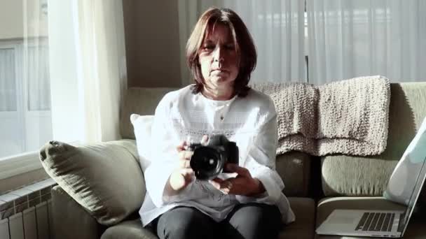 白いシャツに身を包んだ茶色の髪の写真家の女性は 自宅のソファから快適にカメラを構えています 彼女は社会的孤立に準拠している — ストック動画