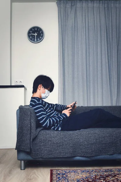 Evde küçük Asyalı bir çocuk, ağız maskesi takıyor ve tablet üzerinde oyun oynuyor (konsept ev karantinası)
