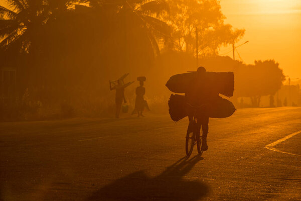 Мозамбикский велосипедист везет на рынок большие мешки угля на рассвете, вдоль взлетно-посадочной полосы. Другие пассажиры перевозят предметы на заднем плане. Нампула-Таун
 