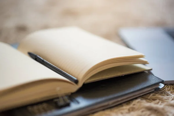 An arrangement of an open black journal and pen with a laptop on a rug on the floor, with warm light coming from above