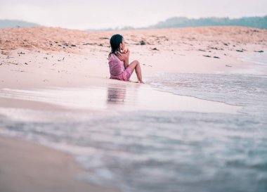 Filipinli zavallı kız denize bakıyor ve pembe elbiseleriyle kumda oturuyor.