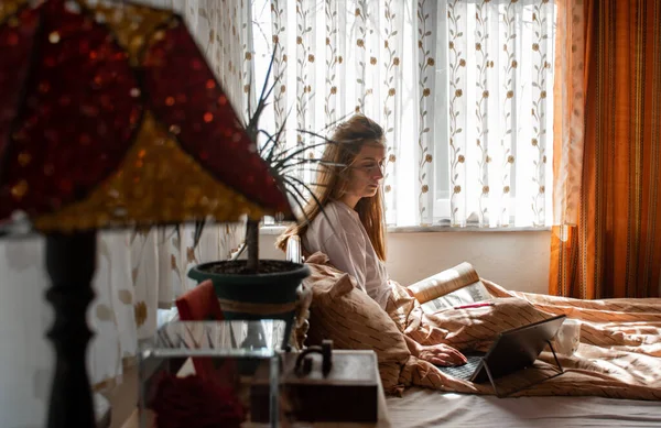 Genç bir bayan yatakta kitap okuyor ve sabahın erken saatlerinde tablet bilgisayardan hafif güneş ışığıyla bir şeyi kontrol ediyor.