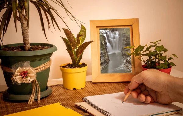 Bir adam defterine bir çizim çiziyor, çiçek saksılarının önündeki masanın üzerinde çalışıyor ve çerçeveli manzara resmi, yakın plan.. 