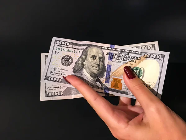 Gri bir arkaplanda iki tane 100 Dolarlık banknot tutan bir kadının eli.