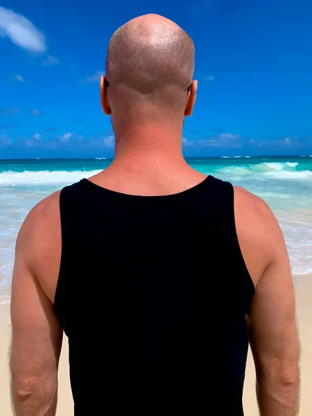 European hairless bearded man male guy standing on the beach in black sleeveless shirt singlet with empty space for text or print on ocean sea background. View from back