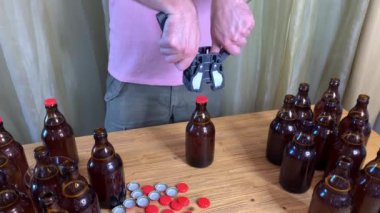 Evde bira demleyen bir adam, kahverengi cam bira şişelerini kapar. Ahşap masa üzerinde kırmızı kapaklı plastik kapaklı. Yatay HD stok videosunu kapat.