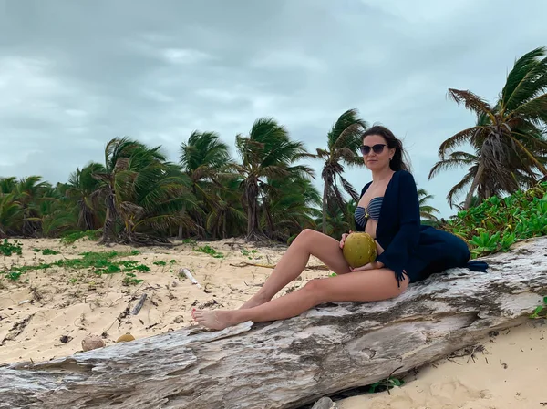 彼女の手にココナッツと若い暗い髪のヨーロッパの女の子は 背景に曇った空とココナッツヤシの木とドミニカ共和国の白い砂浜カリブ海の上に倒木に座っています — ストック写真