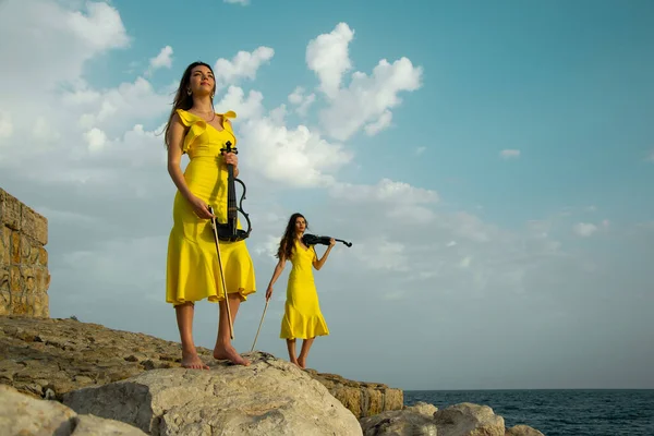 黄色のコンサートドレスを着た2人の美しい双子の姉妹のヴァイオリニストは トルコのアンタルヤで地中海の岩の上で電気バイオリンを演奏しています 背景に海と曇りの空 ストック画像 ロイヤリティフリーのストック写真