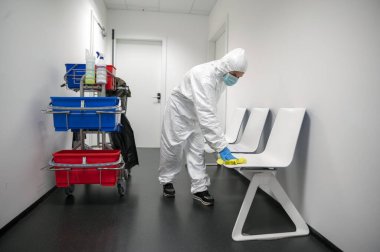 Corona virüsü ya da Covid-19 'u uyarın. Koruyucu takım elbiseli, maskeli ve eldivenli bir temizlikçi hastane, klinik ya da bilinçli bir yerin bekleme odasını temizler, dezenfekte eder ve sterilize eder. Sandalye temizliği.