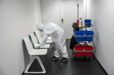 Corona virüsü ya da Covid-19 'u uyarın. Koruyucu takım elbiseli, maskeli ve eldivenli bir temizlikçi hastane, klinik ya da bilinçli bir yerin bekleme odasını temizler, dezenfekte eder ve sterilize eder. Sandalye temizliği.