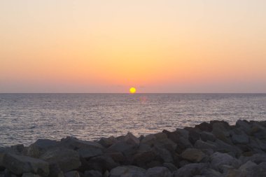 Denizin üzerinde gün batımı, ön planda taşlar.