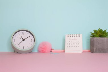 Durgun bir hayatın yan görüntüsü. Pembe kabarık kalem, saat ve takvim pembe bir masada. Arkasında turkuaz duvar var.