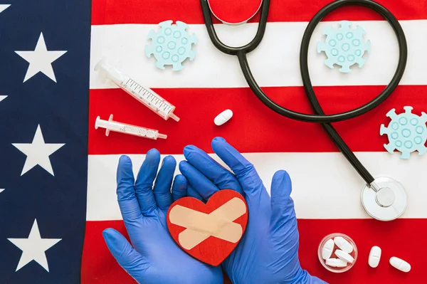 Medical concept flat lay of a medic\'s hand holding a heart, pills, stethoscope and vaccines near to cardboard coronavirus on American flag as background and copy space